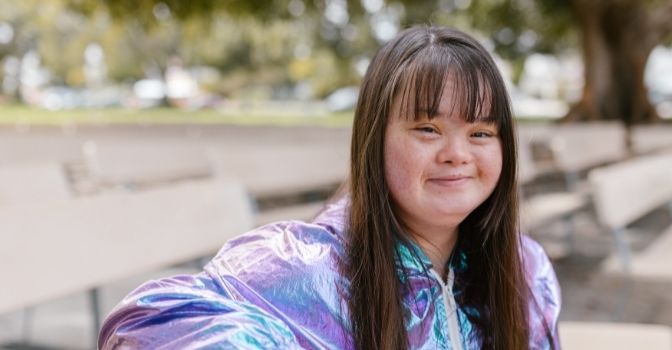 A lady is sitting on a bench in a park looking at the camera with a smile on her face.