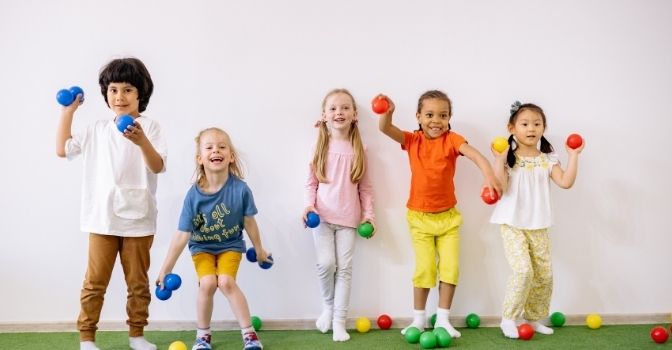 Five children are all smiling facing the camera and picking up and throwing a variety of coloured balls.
