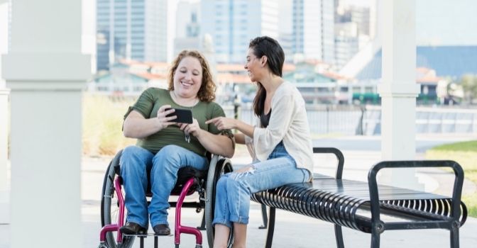 A woman in a wheelchair is showing her friend who is sitting on a park bench next to her what is on her phone. Both women are smiling happily.