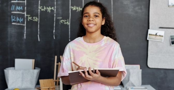 A girl is standing in front of a class room chalkboard smiling at the camera.