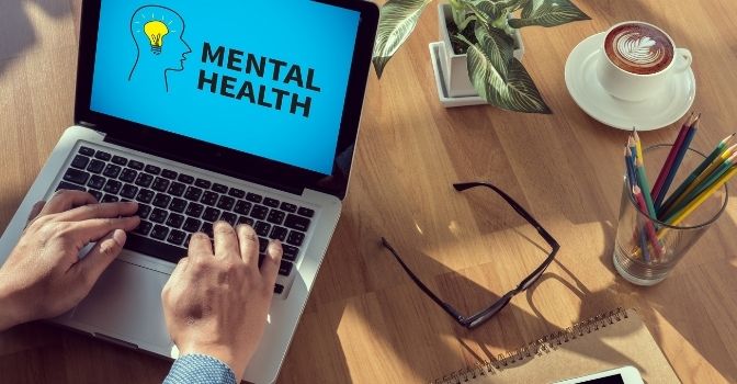 A laptop is on a desk with the words 'Mental Health' on the screen on a blue backdrop.