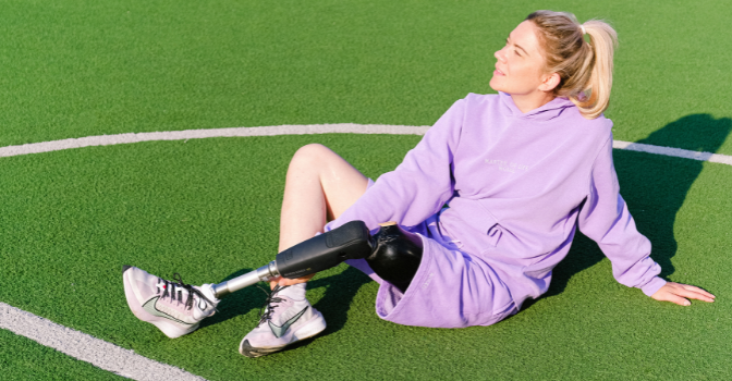 A woman para athlete sits in the sun in purple workout clothing on a green sports field.