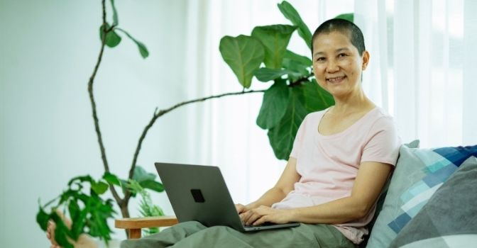 A woman is sitting on her lounge with her feet up, a laptop in her lap and is smiling toward to the camera.
