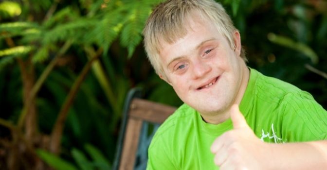 A boy with Down Syndrome in a green shirt is looking at the camera smiling with a thumbs up.