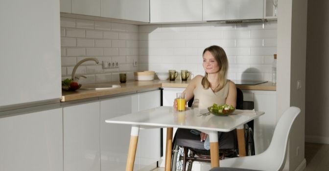 A woman in a wheelchair is sitting at her kitchen table holding a glass of juice and smiling.