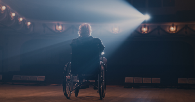 A man in a wheelchair on stage faces the crowd with stage camera lights shining on him.