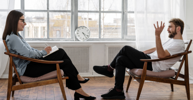 A man sits in a chair on the right and a woman sits in a chair on the left facing each other in front of a window. the man is talking and gesturing and the woman is holding a notebook and pen.