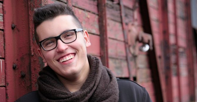 Man with glasses leans against a wall outside and smiles