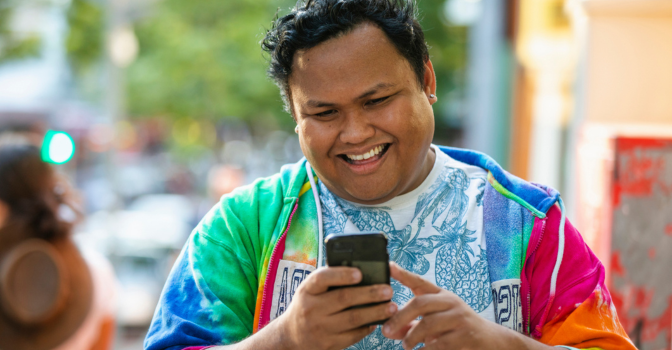 Young adult walking a down a street smiling at their phone, wearing a rainbow coloured shirt.