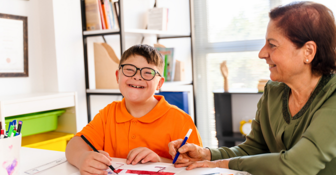 A young boy with disability, writing in class with help from his teacher.