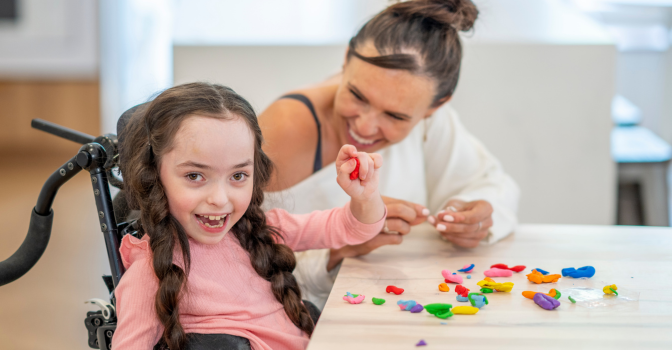 A female social worker sits with a young girl battling Cerebral Palsy, as they work together on some basic skills. The worker is dressed casually and the little girl is seated in her wheelchair pulled up to the table as they play with Playdoh together.