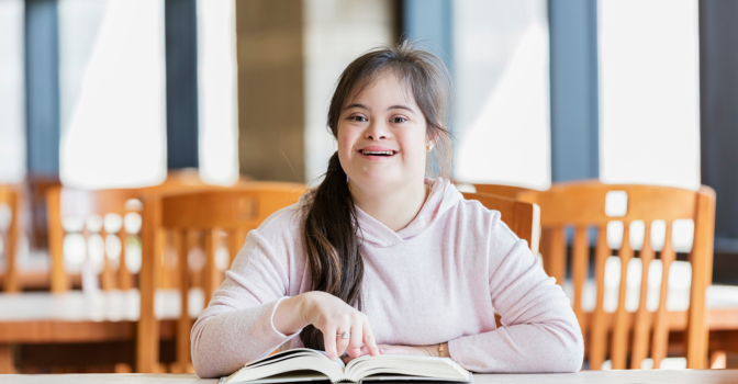A teenage girl with down syndrome, sitting at a table in the library, reading a book.