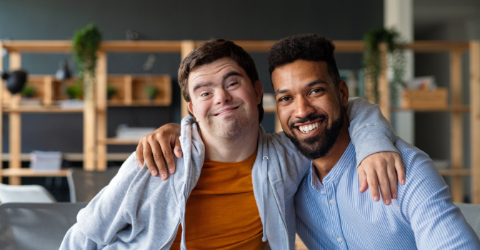 a young man with a disability embracing his carer, both smiling and looking at the camera