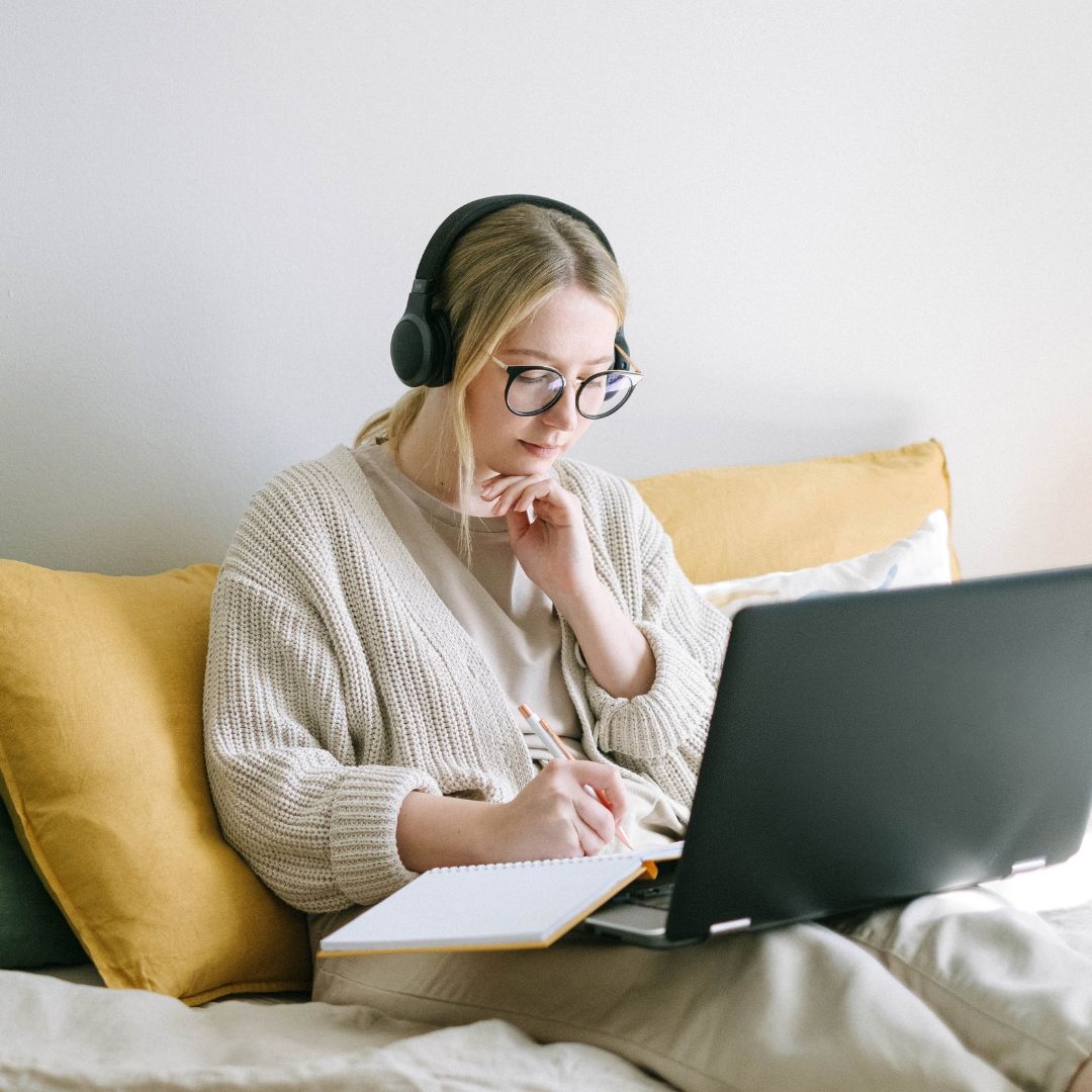 Woman sitting on a lounge with a laptop on her lap, a notebook and headphones.