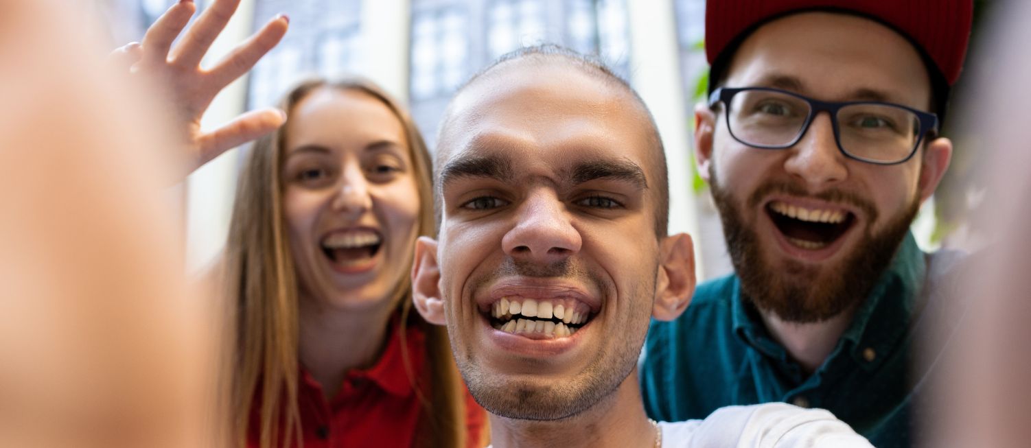 Man holding camera to take a selfie of himself and a man and a woman who are standing behind him smiling