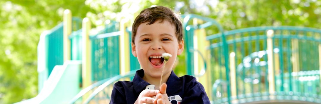 Young boy standing in front of a playground holding a flower