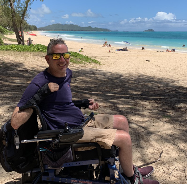 Lindsay sitting in his wheelchair at a beautiful beach with sparkling blue water. He is smiling and wearing sunglasses.