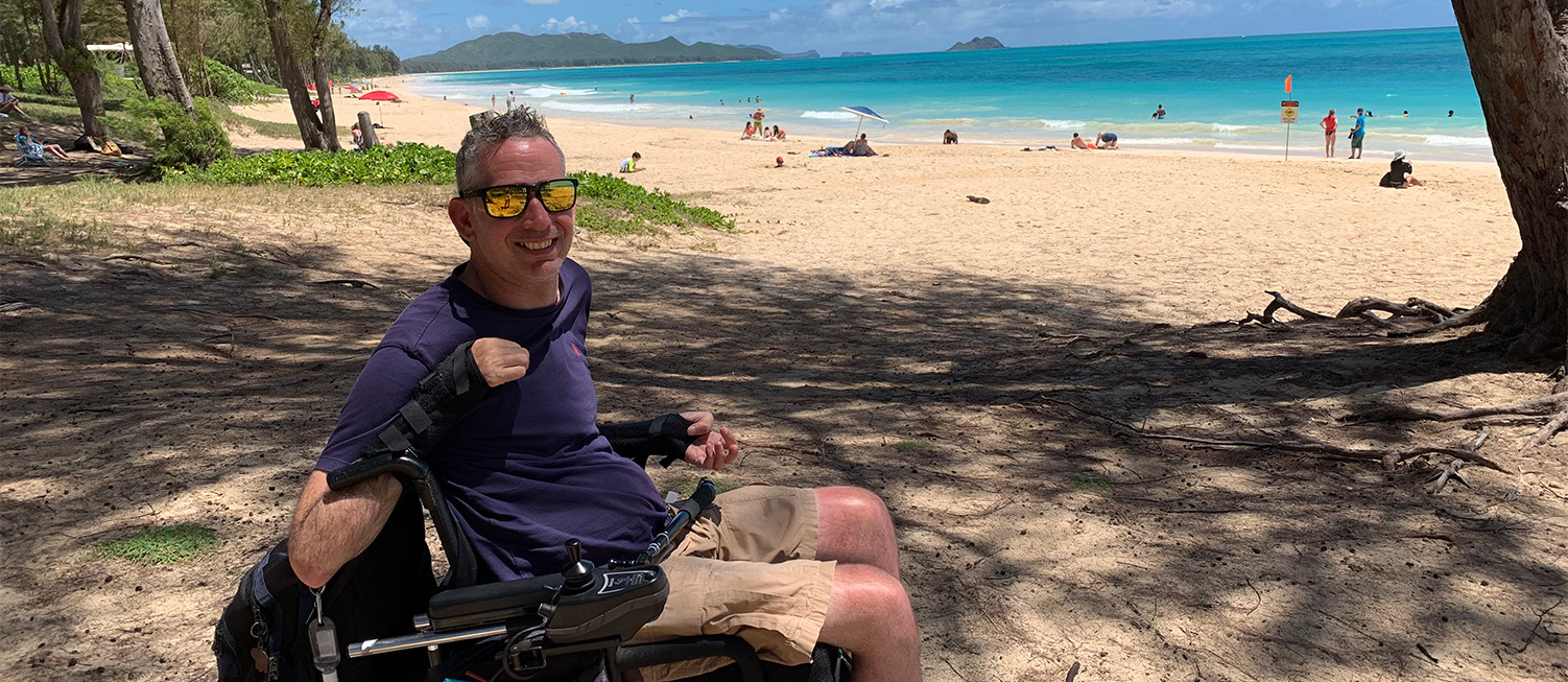 Lindsay sitting in his wheelchair at a beautiful beach with sparkling blue water. He is smiling and wearing sunglasses.