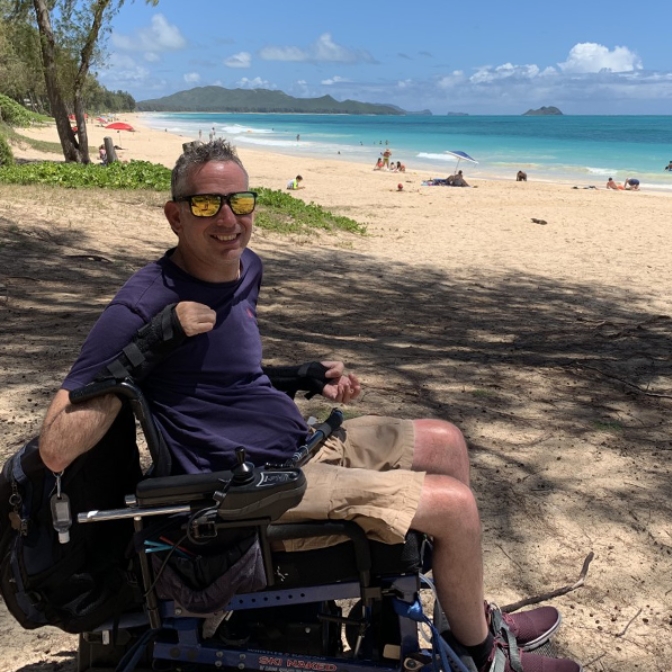 Lindsay is on the beach in his wheelchair smiling. It is a beautiful sunny day in the background.