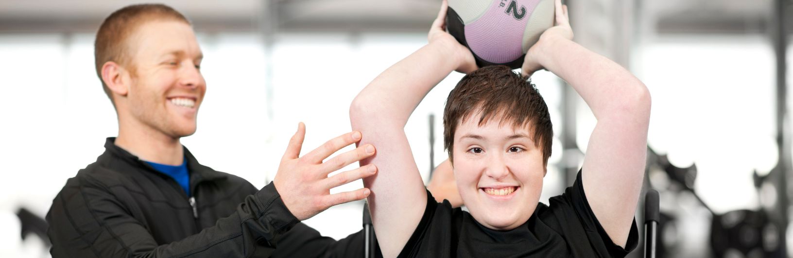 Young woman in a wheelchair holding a small ball over her head with a personal trainer at her side