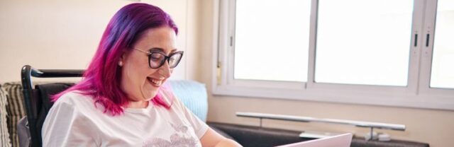 Woman with bright pink hair and black glasses in an electric wheelchair with a laptop on her lap