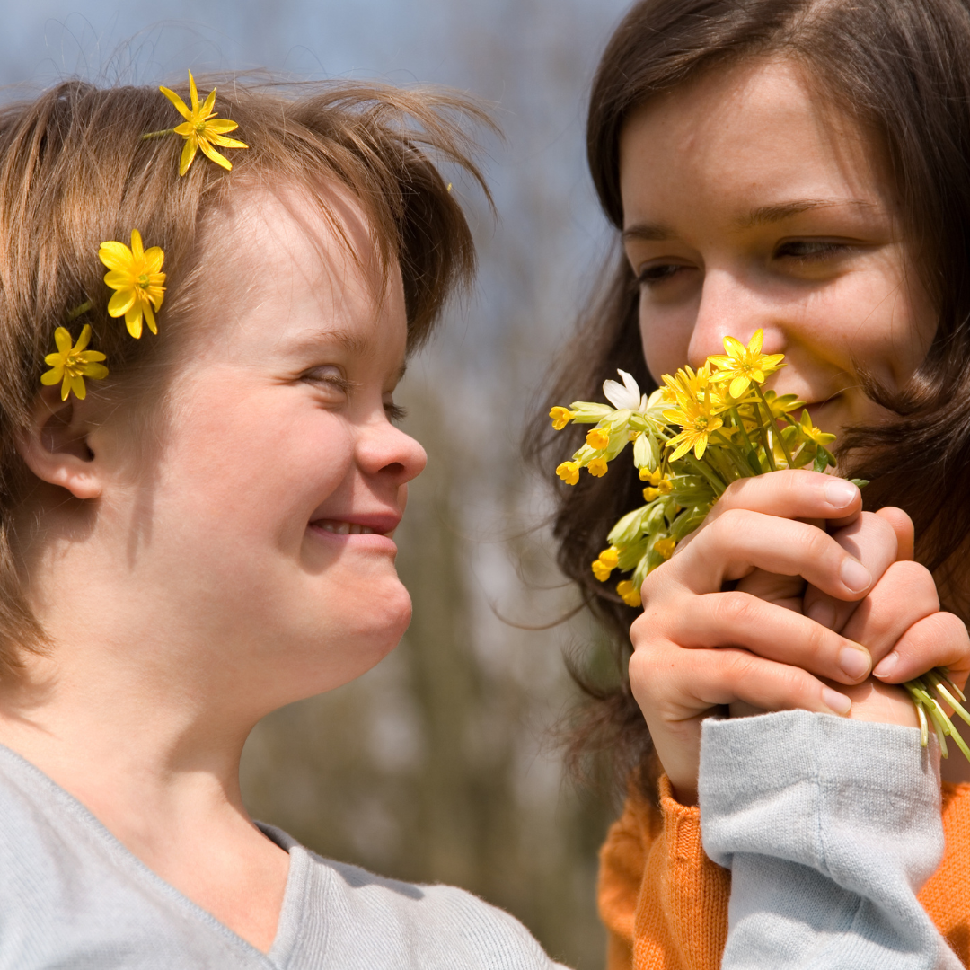 Girl holding a small posy of yellow wildflowers to the nose of her friend.