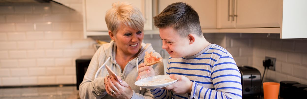 Woman standing in a kitchen showing a young man her phone while he eats toast